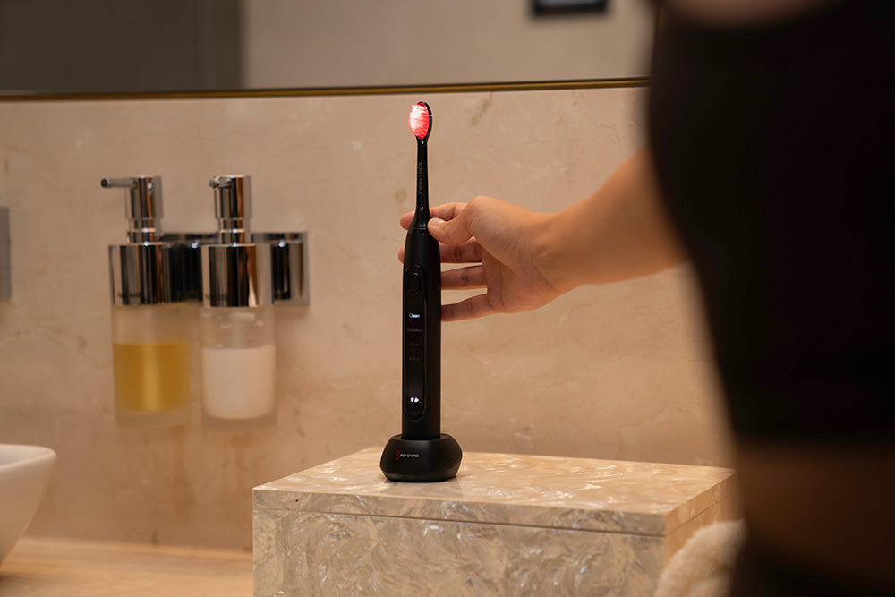 Person holding a black electric toothbrush with a red light on a bathroom counter.